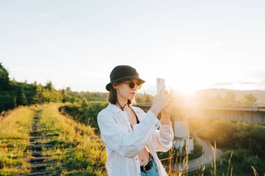 Şık, hafif elbiseli çekici bir bayan, eski bir köprüde günbatımının arka planında duran akıllı bir telefonla fotoğraf çekiyor. Kadın turist köprü arkasında güzel manzaralar çekiyor