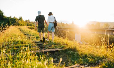 Genç çift eski demiryolu köprüsünde güneşin arka planında yürüyor ve şık yaz kıyafetleri giyip gülümsüyor. Genç turistler dağlarda el ele tutuşuyorlar.