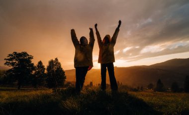 Sarı yağmurluklu iki yürüyüşçü kollarını kaldırarak dağlarda günbatımının arka planında bir çayırda dikiliyorlar. Yağmurda gün batımının arka planında neşeli turistlerin silueti