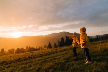 Akşam saatlerinde dağlarda, gün batımında, yağmur altında yürüyüş yapan iki yürüyüşçü. Sarı yağmurluklu iki insan gün batımında yağmurun altında dağlardaki bir çayırda durup manzaranın güzelliğine bakarlar..