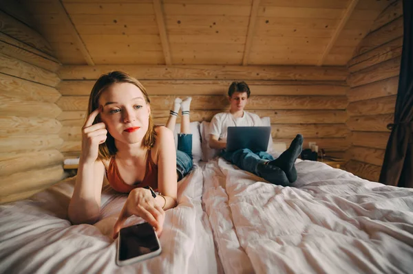Unhappy girl with a smartphone lies on the bed with a smartphone, touches the temple and looks away with an evil face on the background of a man working on a laptop on a bed. Quarrel of a young couple