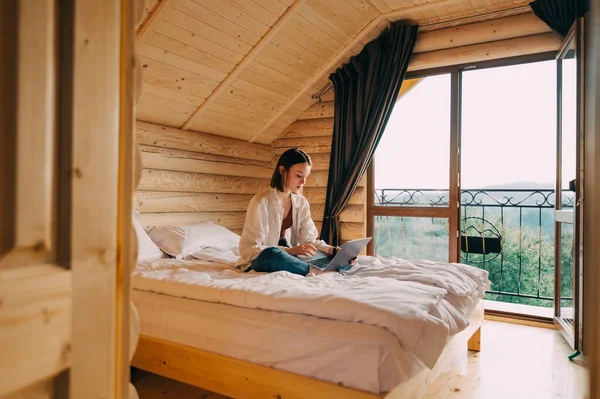 Woman freelancer in casual clothes working remotely on laptop in country wooden apartment. Female freelancer working online on laptop in hotel bed. Work online. Quarantine.