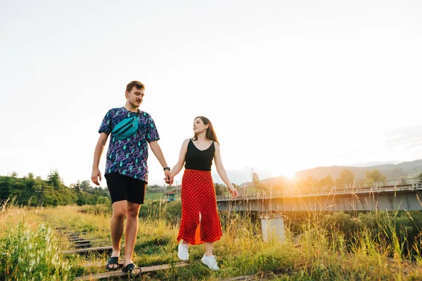 Beautiful couple of young people walking on a viaduct on a background of sunset holding hands. A positive couple of tourists walking on the old railway bridge.