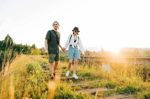 young couple walks on the old railway bridge against the backdrop of the sunrise and smiles, wearing stylish summer clothes. Young tourists go holding hands on a viaduct in the mountains