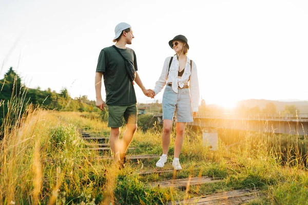 young couple walks on the old railway bridge against the backdrop of the sunrise and smiles, wearing stylish summer clothes. Young tourists go holding hands on a viaduct in the mountains
