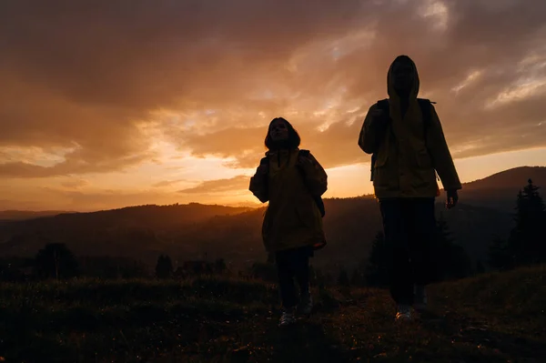 Silhouette of two female hikers climbing mountains in rainy weather on a background of sunset with backpacks on their backs. Two hikers on a hiking trip on a background of mountains at sunset.