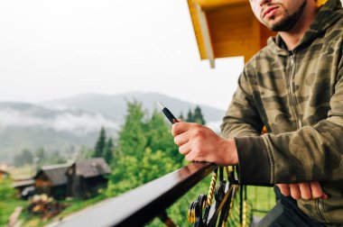 Kapatın. Elektron sigaralı bir erkek el. Bir adamın elinde tütün ısıtan bir cihaz bir dağ dairesinin balkonunda duruyor ve sigara içiyor..