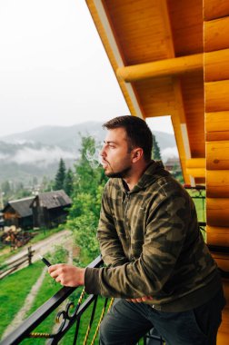 Yetişkin sakallı adam, dağlarda bir apartmanın balkonunda tütünle birlikte elektronik sigara içiyor ve manzaraya bakıyor..