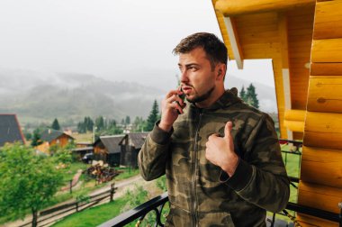 Ciddi sakallı bir adam bir kır evinin balkonunda duruyor ve telefonla konuşuyor. Arkasında ciddi bir yüz olan güzel bir dağ manzarası var..