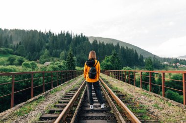 Hippi kadın şık giysiler içinde sırtı dağlarda bir demiryolu köprüsünün raylarında dikiliyor, arkadan bakıyor. Yürüyüş yapan kadın yürüyüşçü dağlarda bir piste gider. Karpatlar