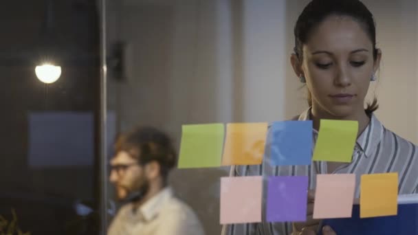 Jeune femme travaillant au bureau et collant des notes sur un mur de verre — Video