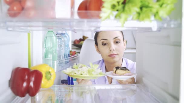 妇女打开冰箱和选择最好的食物为她的饮食 她正在比较健康的蔬菜和美味的甜点 她选择不健康的零食 — 图库视频影像