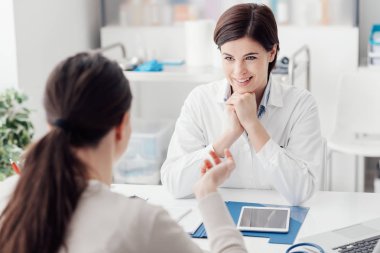 Ofiste çalışan ve hastayı dinleyen doktor semptomlarını, sağlık hizmetlerini ve yardım kavramlarını açıklıyor.