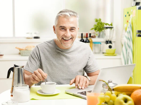 家庭で健康的な朝食を持つ成熟した男の笑みを浮かべて 彼は台所のテーブルに座ってコーヒーを飲んで 彼のラップトップを接続します — ストック写真