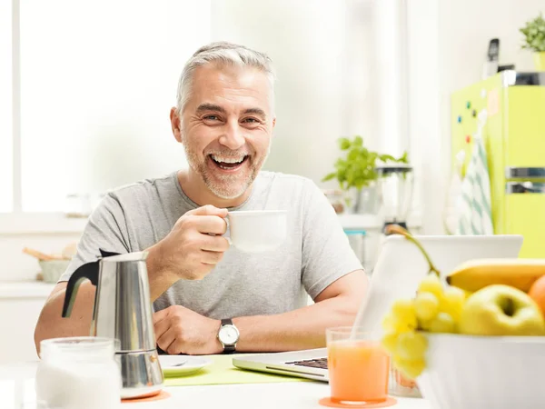 微笑成熟的人在家里吃着健康的早餐 他坐在餐桌旁 喝着咖啡 用笔记本电脑连接 — 图库照片