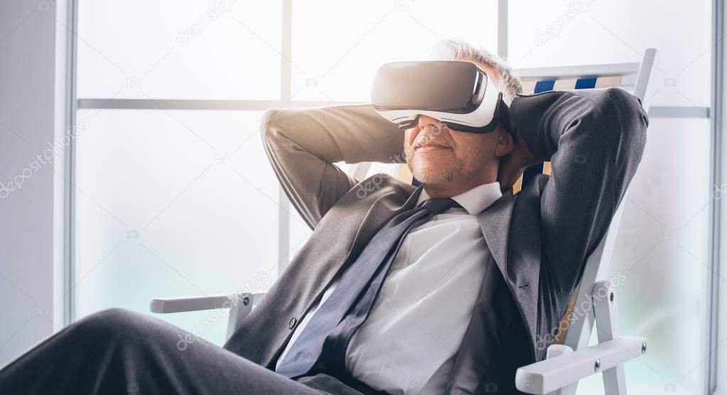 Businessman wearing a VR headset and enjoying a virtual vacation, he is relaxing on a deckchair and smiling; technology and virtual reality concept