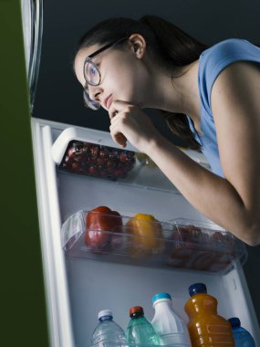 Genç kadın bir yemek hazırlama ve çene üzerinde el ile düşündüğünü yiyecek buzdolabında, arama