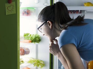 Genç kadın bir yemek hazırlama ve çene üzerinde el ile düşündüğünü yiyecek buzdolabında, arama
