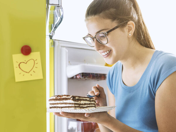 Hungry young woman taking a delicious dessert from the fridge and having a snack, diet fail concept