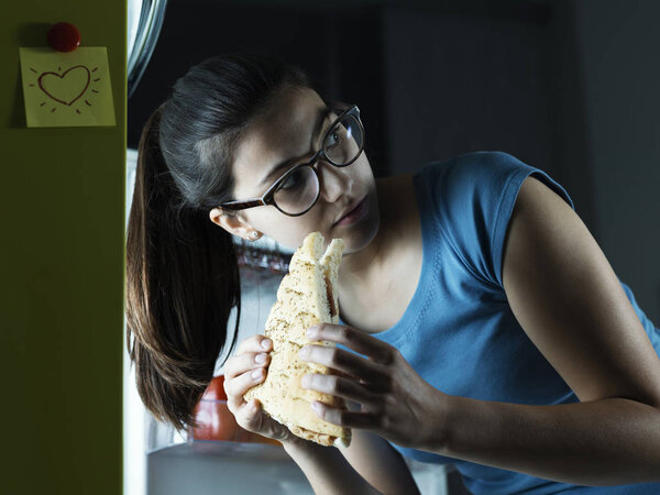 Young woman having a late night snack, she is taking a sandwich fron the fridge and eating it, diet fail concept