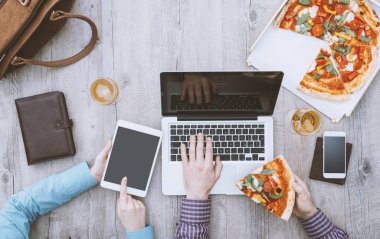 Ofis çalışanları ofiste yemek molası veriyorlar, pizza yiyorlar, dizüstü bilgisayar ve dijital tablet kullanıyorlar, düz konum alıyorlar.