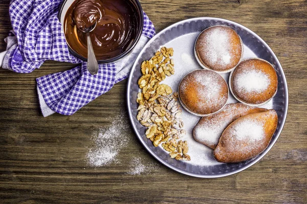 Delicious fresh buns and walnuts in powdered sugar in a metal bowl. Hot chocolate, cooked in a saucepan for buns. Wooden table. Rustic style.