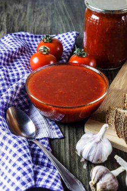 Bir cam kasede salça. Bir kavanoz içinde domates sosu. Olgun domates ve sarımsak. Damalı havlu ve kaşık. Dilimlenmiş ekmek.