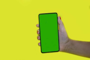 Elinde siyah bir akıllı telefon var, dikey, yeşil ekran, krom anahtar, sarı arka planda..