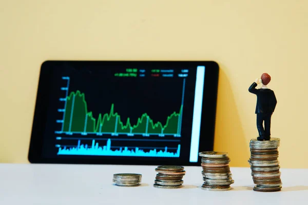İş adamı ve finansal büyüme konsepti, arka arkaya madeni paralar ve tablet ekrandaki borsa grafiği bulanık, bozuk paraların üzerinde duran minyatür işadamı.