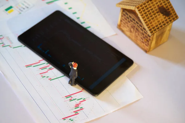 Beyaz kağıt üzerinde borsa grafik verileri olan minyatür işadamı ve bulanık tablet ile bulanık ahşap ev, iş yeri veya borsa grafik analizi kavramı