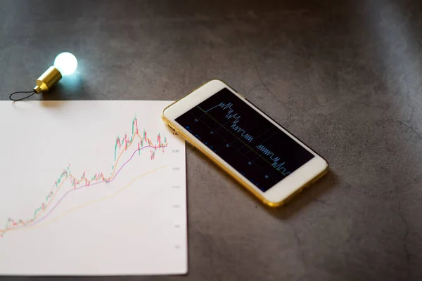 Yeni borsa grafik grafik analizi fikri, akıllı telefon ekranında bulanık ampul ışığı fikri sembollü borsa grafiği, beyaz kağıt üzerinde bulanık kalem ve borsa grafiği.