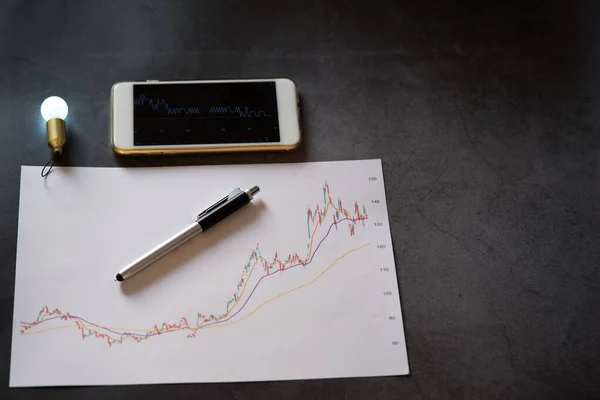 Yeni iş finansal analizi fikri veya borsa grafik analizi konsepti, akıllı telefon ekranında bulanık ampul ışığı fikri, bulanık kalem ve beyaz kağıt üzerinde borsa grafiği