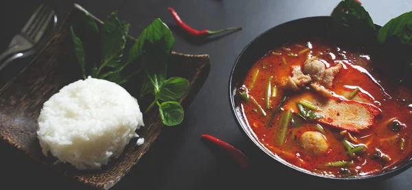 Panoramic banner for web, Thai food background concept. Dish of Thailand cuisine. Tom yum pork soup in black dish, rice in wooden plate on table dark background 2