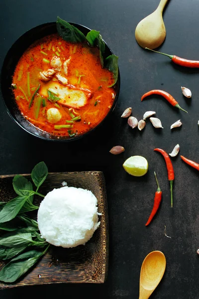 Thai food vertical background concept. Dish of Thailand cuisine. Tom yum pork soup in black dish, rice in wooden plate, wooden ladle and spoon, chilis, garlic on table dark background