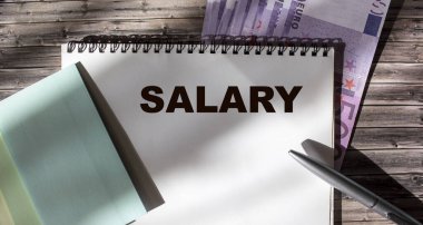 Not defteri, kalem, çıkartma ve ahşap masadaki euro banknotlarına ilişkin maaş metni