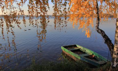 Gölde teknesi olan renkli bir sonbahar manzarası. Boş ahşap tekneli kır gölü hattı Eylül sarısı ağaçlar ve gün batımı turuncu güneş ışığı parlıyor.