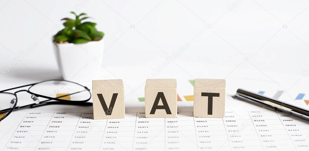 VAT word concept written on wooden blocks, cubes on a light table with flower ,pen and glasses on chart background