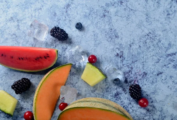 Taze kesilmiş kavun, karpuz, böğürtlen ve buz küpleri mavi taştan arka planda. Boşluk ve metin alanını kopyala