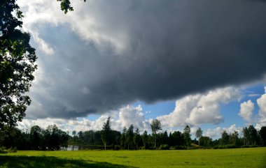 Yaz boyunca kara fırtına bulutları, Letonya 'nın ön planında ağaçlar ve çayırlar olan manzara. Düşük bulutlar