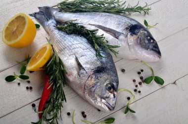 Beyaz ahşap arka planda limonlu, soğanlı ve biberiyeli iki taze Dorado balığı.