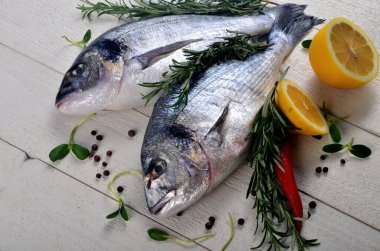 Beyaz ahşap arka planda limonlu, soğanlı ve biberiyeli iki taze Dorado balığı.