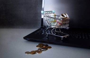 Karanlık bir arka planda dizüstü bilgisayarında parası olan küçük bir alışveriş arabası. İş ve mali konsept.