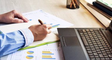 Dizüstü bilgisayarın arka planında kadın eli ve iş grafikleri var. Kız masadaki ev ofisinde çalışıyor..