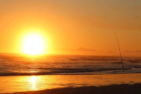 Sunset over the sea, with small waves and clear sky. A fishing rod waiting to get a catch on the right and mountains visible in the distance across the sea. Sun reflecting off the water.