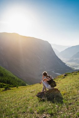 Genç bir kadın, dağların arasındaki güzel bir vadinin arka planında oturuyor. Sırt çantasıyla yürüyüş. Harika bir manzara. Doğayla baş başa.
