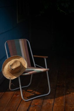 Tahta bir güvertede yeşil asma yapraklarıyla çevrili bir yaz şapkası ve katlanır bir sandalye. Kır evi estetiği. Şehir dışında bir tatil. Sıcak yaz günü.