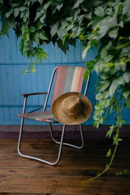 Tahta bir güvertede yeşil asma yapraklarıyla çevrili bir yaz şapkası ve katlanır bir sandalye. Kır evi estetiği. Şehir dışında bir tatil. Sıcak yaz günü.