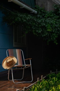 Tahta bir güvertede yeşil asma yapraklarıyla çevrili bir yaz şapkası ve katlanır bir sandalye. Kır evi estetiği. Şehir dışında bir tatil. Sıcak yaz günü.