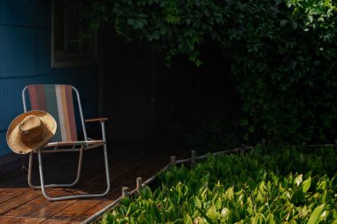 Tahta bir güvertede yeşil asma yapraklarıyla çevrili bir yaz şapkası ve katlanır bir sandalye. Kır evi estetiği. Şehir dışında bir tatil. Sıcak yaz günü.