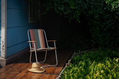 Tahta bir güvertede yeşil asma yapraklarıyla çevrili bir yaz şapkası ve katlanır bir sandalye. Kır evi estetiği. Şehir dışında bir tatil. Sıcak yaz günü.